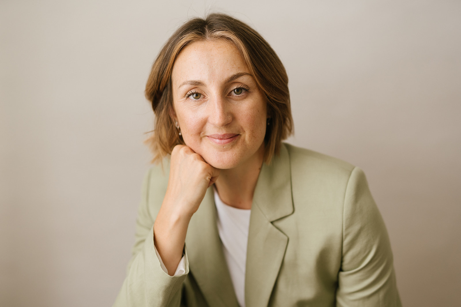 A professional woman with short brown hair wearing a light green blazer and white top, resting her chin on her hand, smiling softly, set against a neutral background.