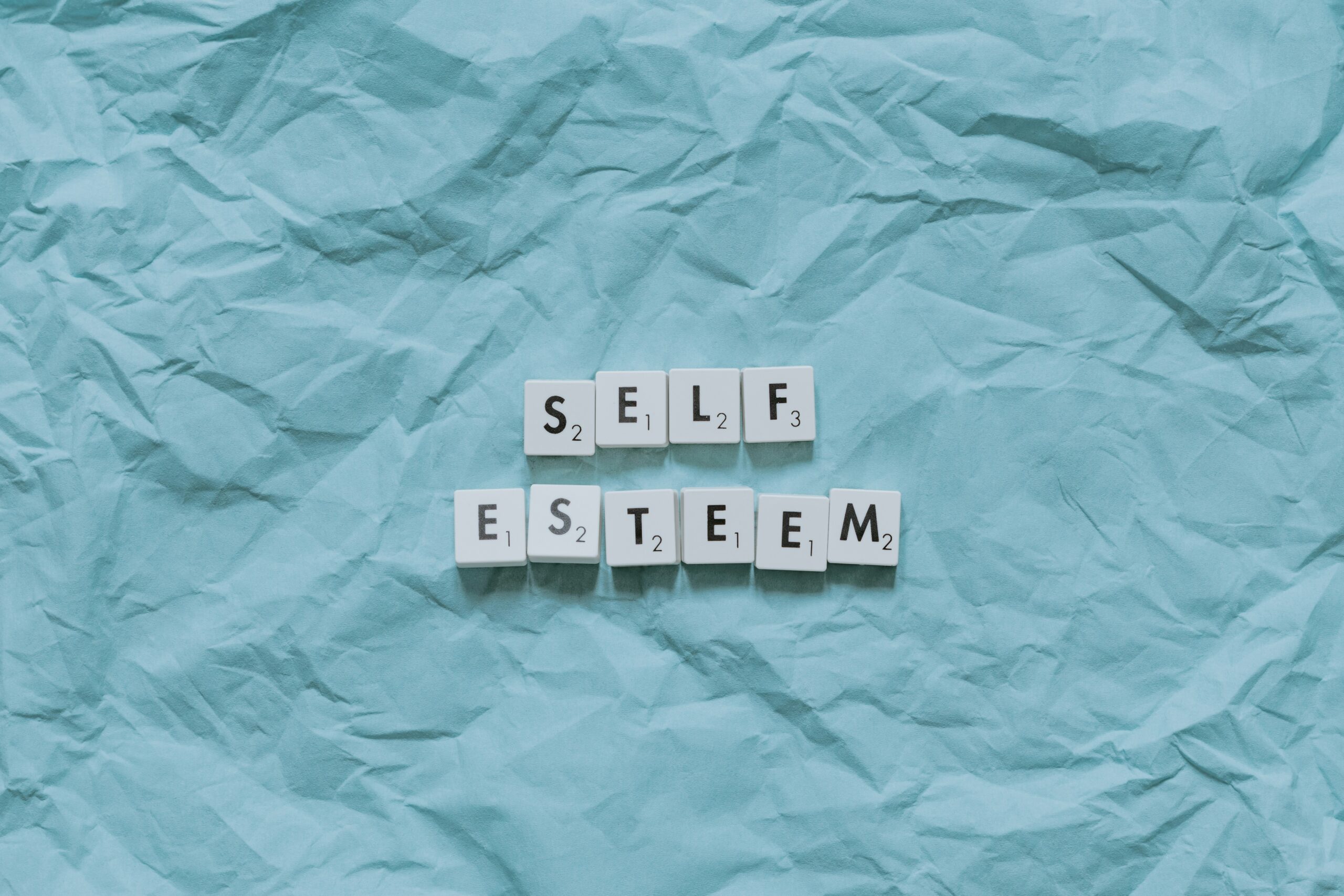 Scrabble tiles spelling out “Self Esteem” on a textured blue background, symbolising the link between energy protection and confidence for professionals managing chronic illness.