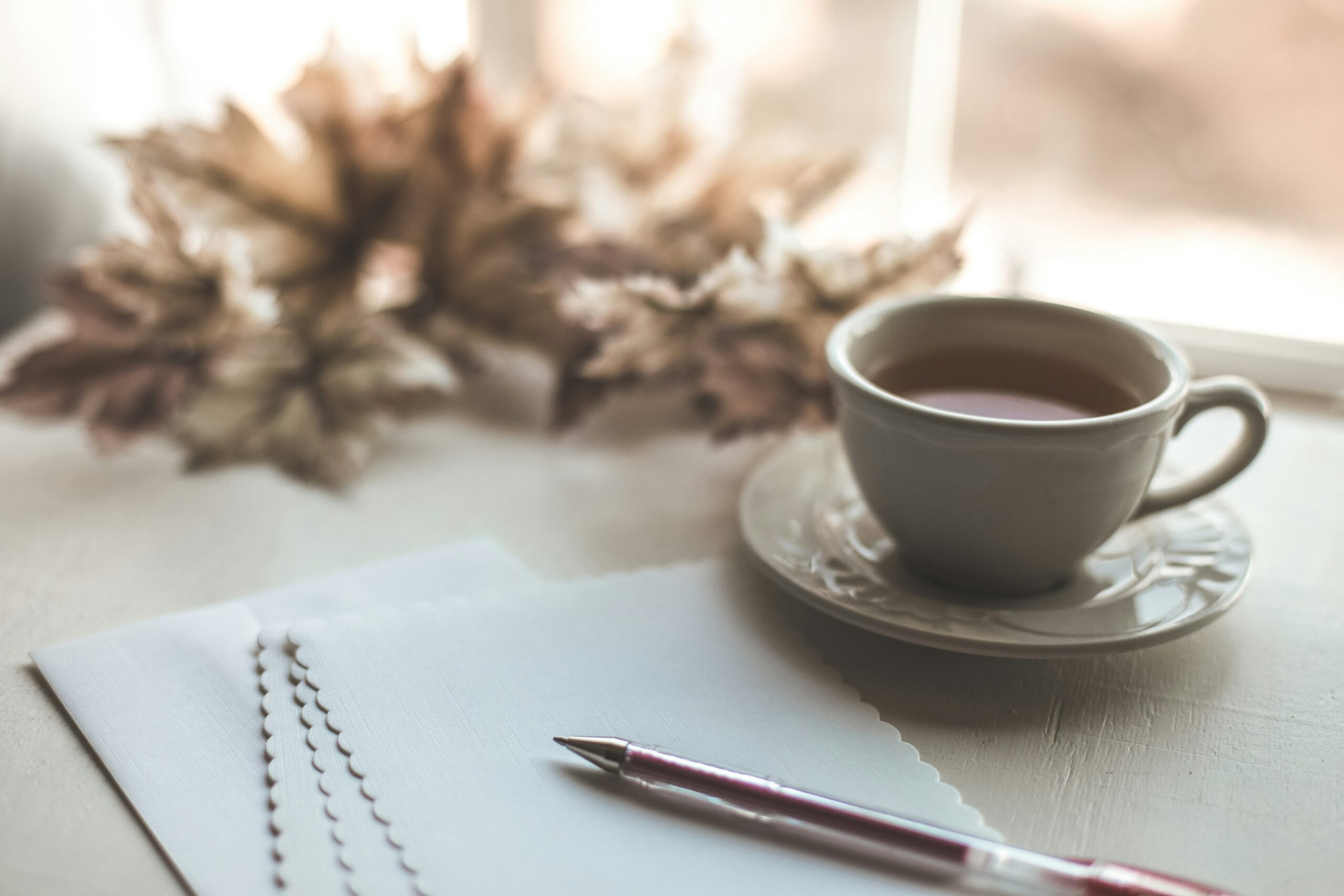A calming scene featuring a cup of tea on a saucer, placed beside blank scalloped-edge stationery and a pink pen, with soft-focus autumn leaves in the background by a sunlit window.