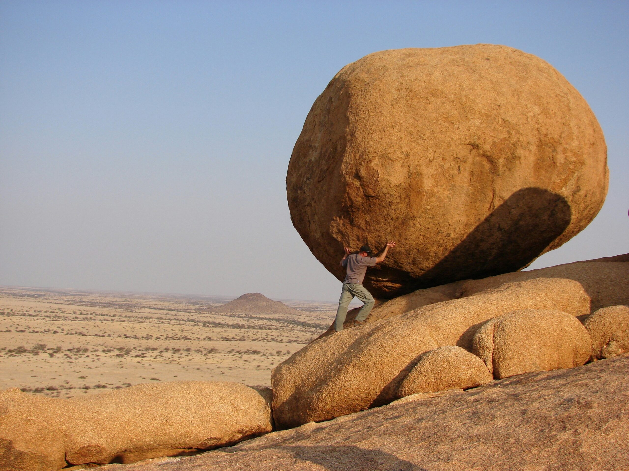 A man pushing a massive boulder uphill in a barren landscape, symbolising the struggle of trying to push through illness at work without proper support or rest.