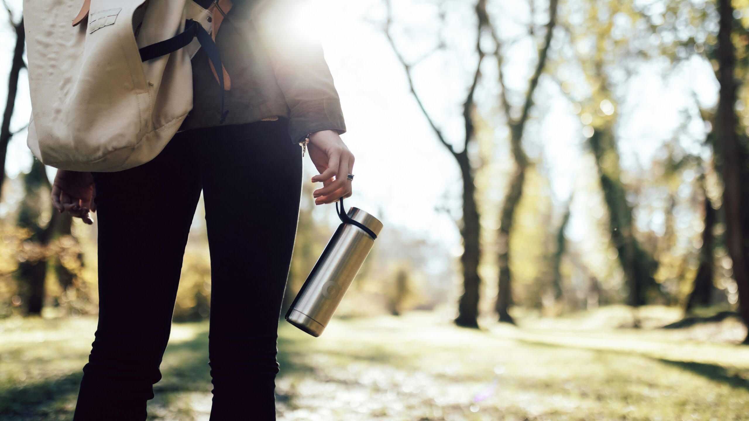 A person holding a reusable water bottle and walking through a sunlit forest, symbolising mindful energy management and the importance of rest, movement and connection to nature for sustainable success.