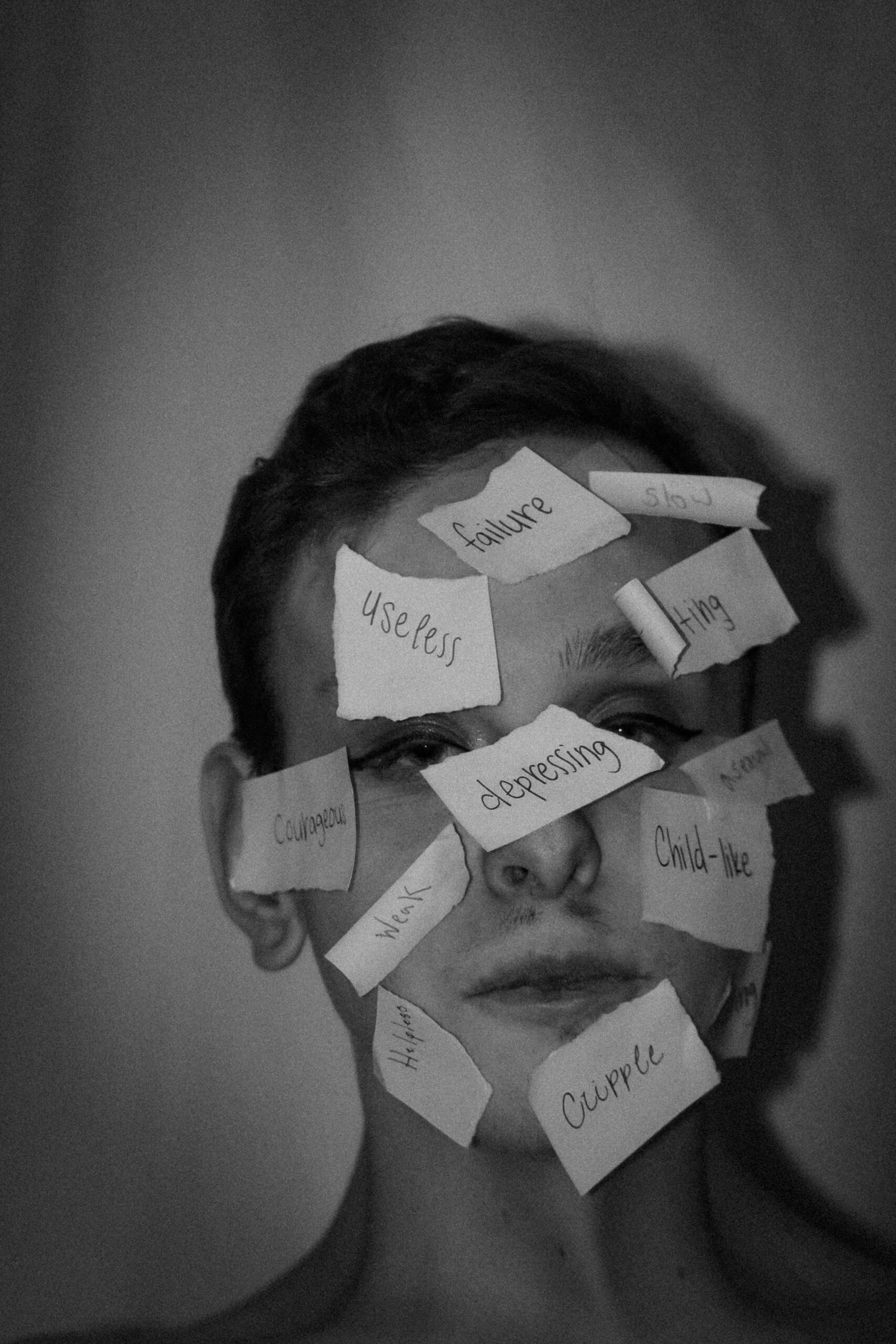 Black and white photo of a person with handwritten labels such as “failure,” “useless,” “depressing” and “child-like” stuck to their face, symbolising the impact of internalised labels and societal judgement on self-worth and identity.