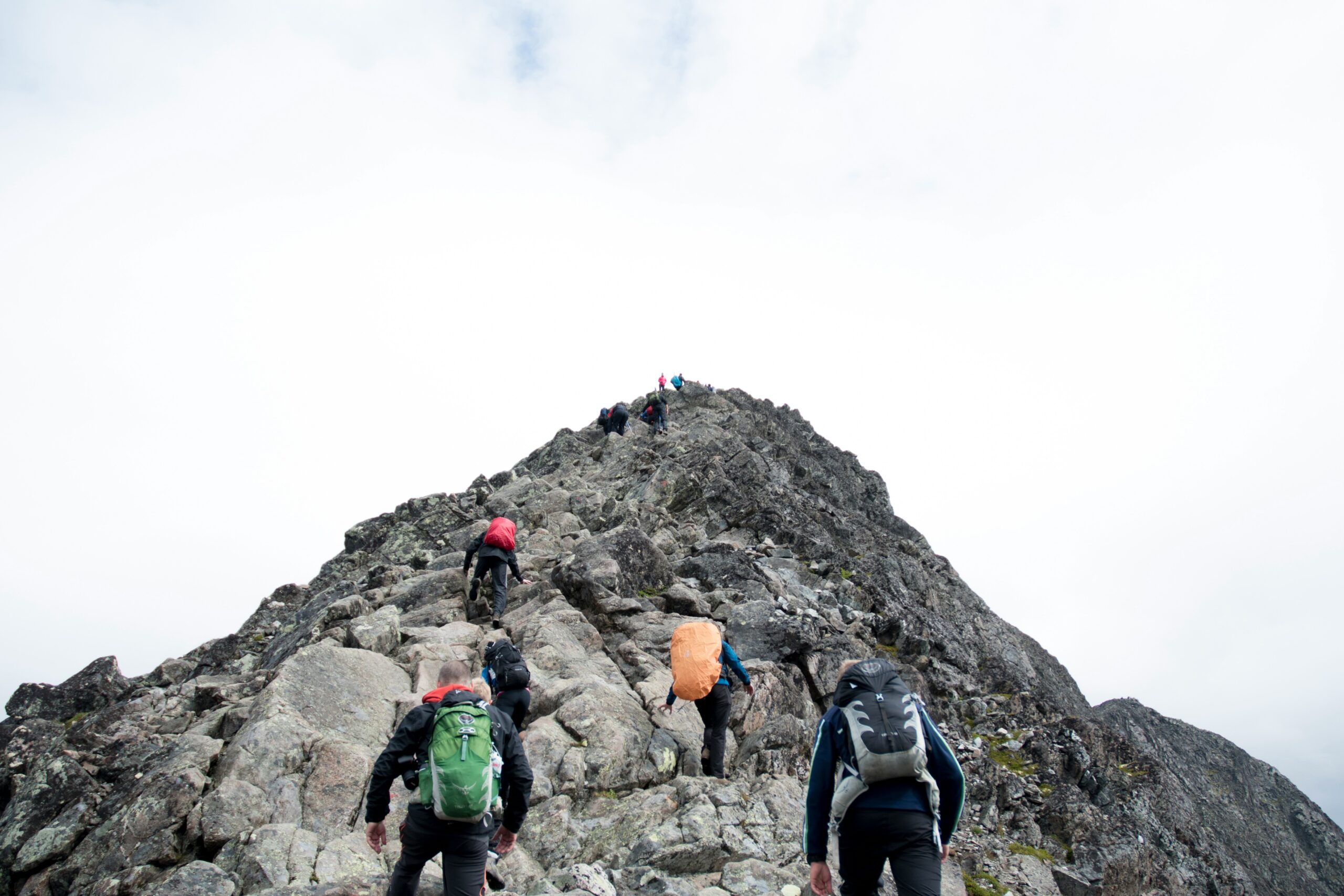 Group of hikers climbing a steep, rocky mountain ridge under an overcast sky, each carrying backpacks in different colours.