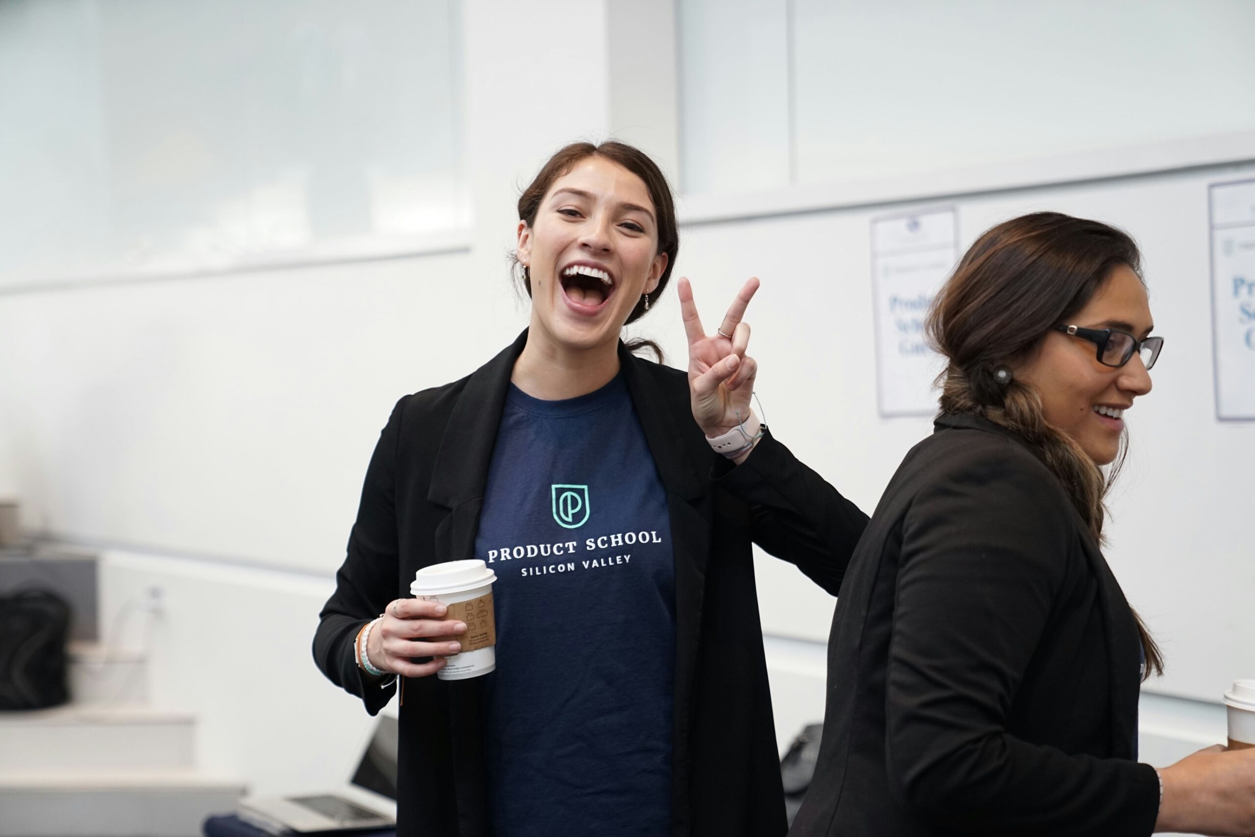 Two women in business casual clothing are smiling and holding takeaway coffee cups in a bright, modern classroom or office setting. One woman, wearing a “Product School Silicon Valley” t-shirt and black blazer, is laughing and flashing a peace sign with her fingers. The other woman is looking down and smiling. Both appear to be enjoying a light-hearted moment during a professional event or workshop.