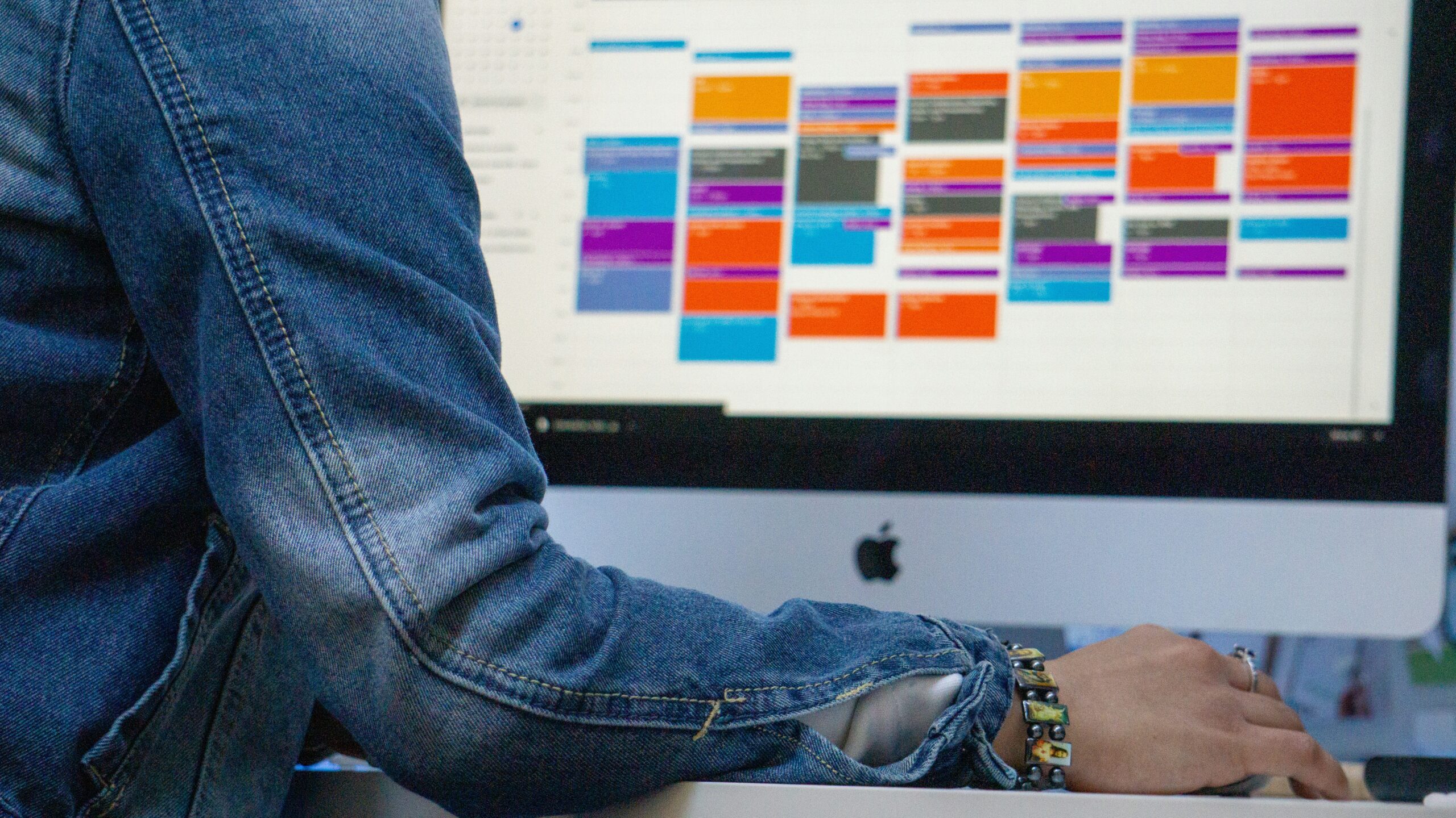 A person in a denim jacket sits at a desk, reviewing a packed digital calendar on a desktop screen — visually representing the invisible energy cost of back-to-back meetings and over-scheduled workdays.