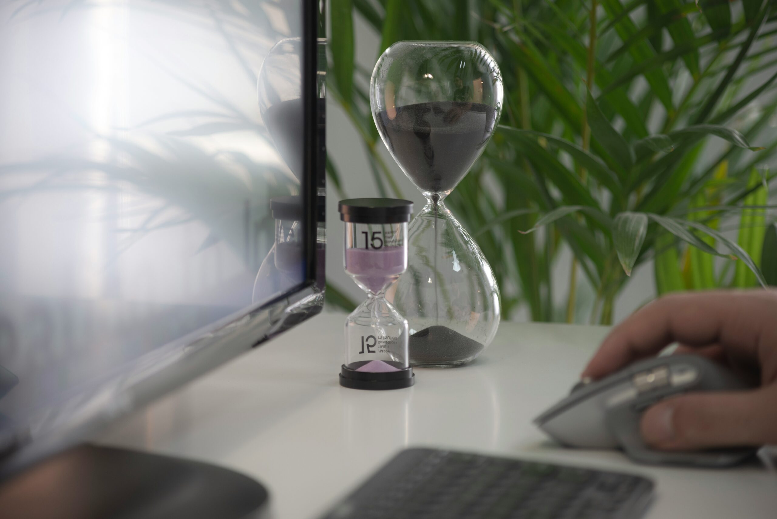 A person using a computer mouse at a desk with two hourglasses in the foreground — symbolising how chronic illness and productivity often require a different relationship with time, pacing and energy management.